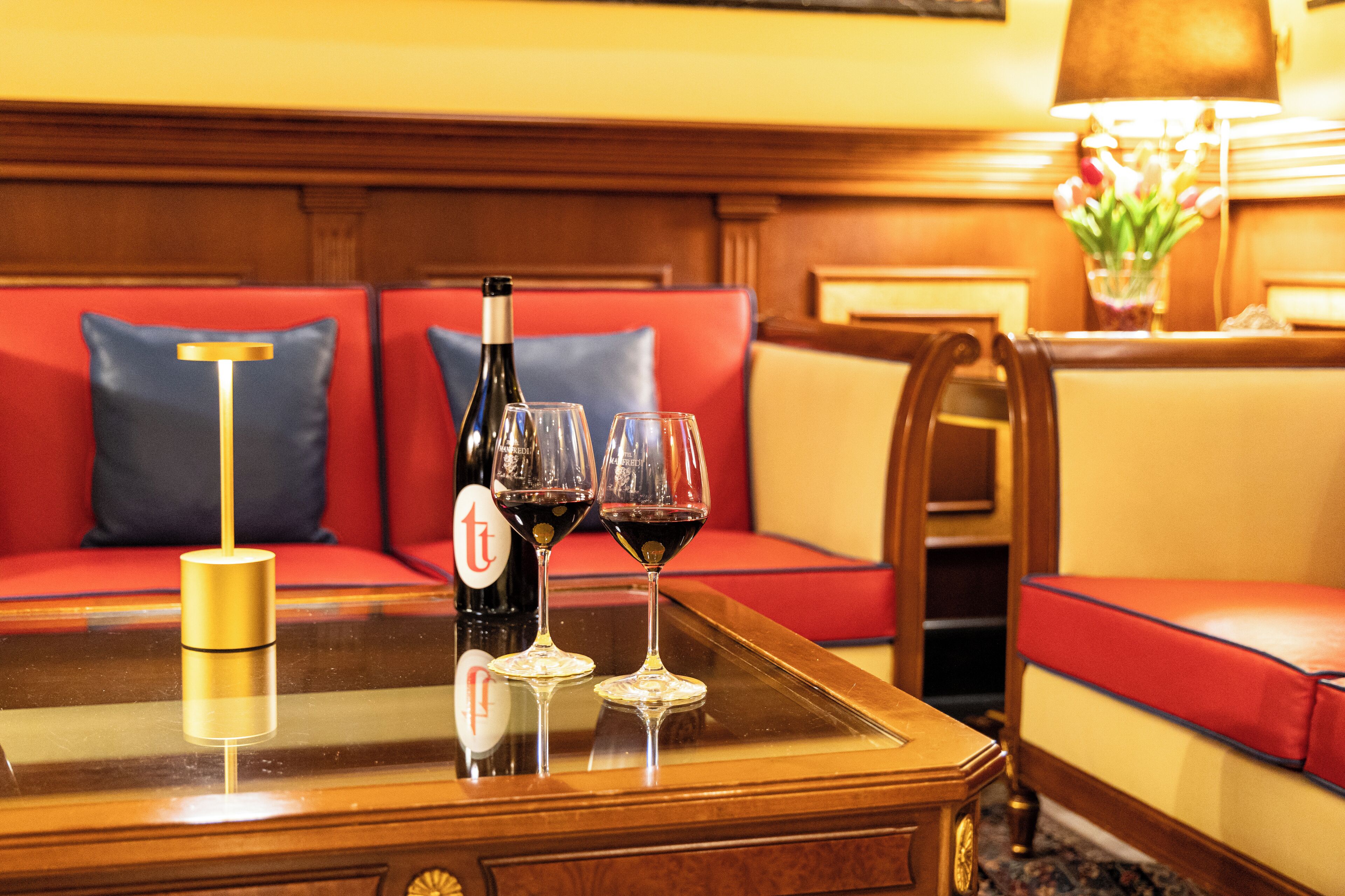 Une chambre d’hôtel avec une table à plateau en verre, une bouteille de vin, deux verres à vin et une lampe.