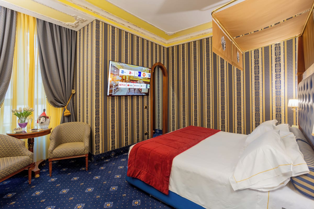A hotel room with a bed, a TV mounted on the wall, a chair, and a small table with a vase and a lamp.