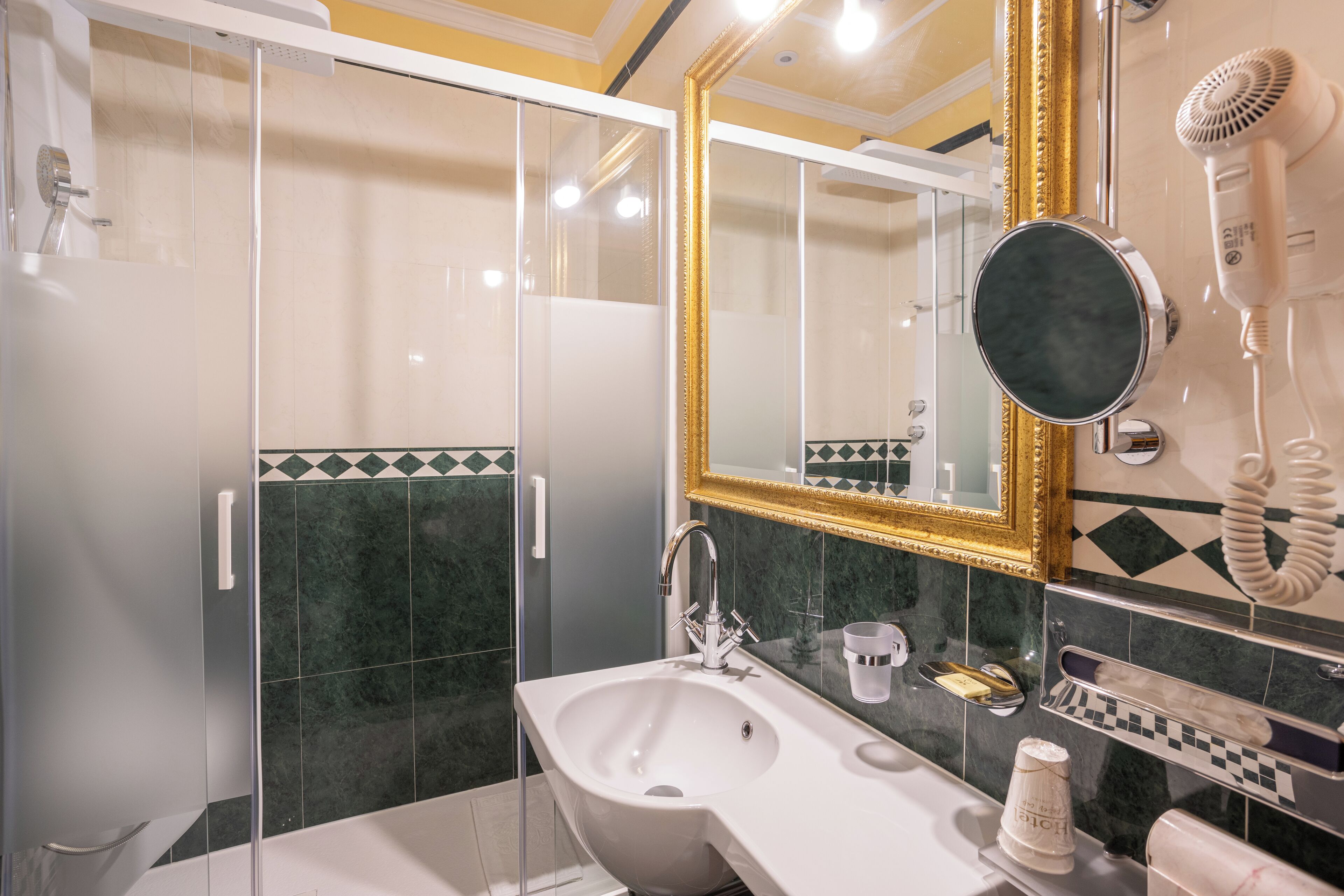 Un bagno moderno con box doccia in vetro, un lavandino bianco, uno specchio grande con cornice dorata e un asciugacapelli fissato alla parete.