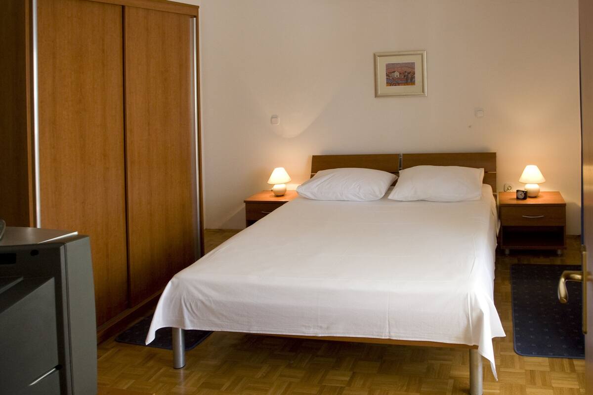 A neatly made bed with white linens, two bedside tables with lamps, a wooden wardrobe, and a small picture on the wall.
