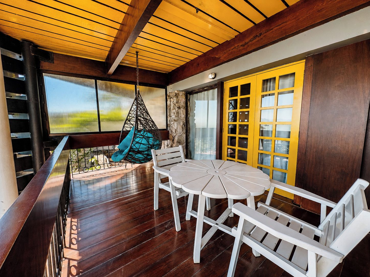 A wooden deck with a hanging chair, a round table, and chairs, and a yellow door with glass panels.