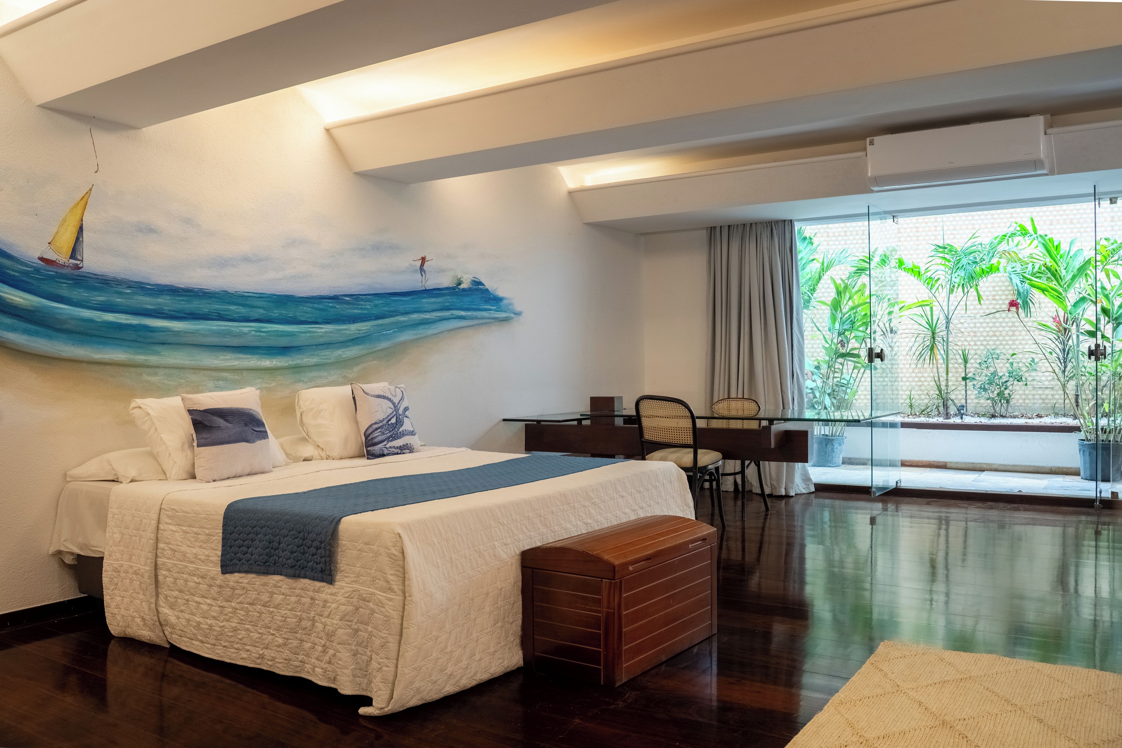 A modern bedroom with a large bed, a desk, and a view of greenery through a window.