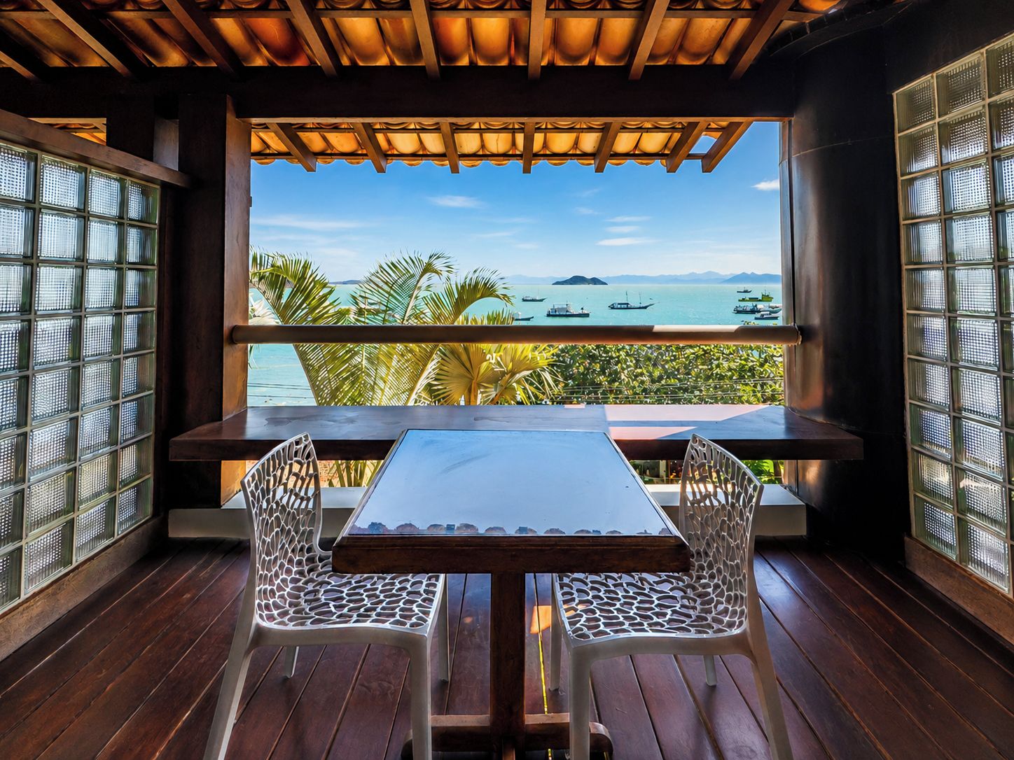 A wooden deck with a table and chairs, overlooking a body of water with boats and palm trees.