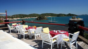 Une terrasse dotée de chaises et de tables blanches, offrant une vue sur une zone côtière avec des bateaux et des collines.