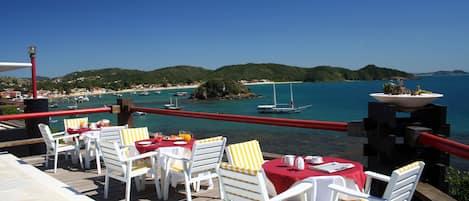 Une terrasse dotée de chaises et de tables blanches, offrant une vue sur une zone côtière avec des bateaux et des collines.