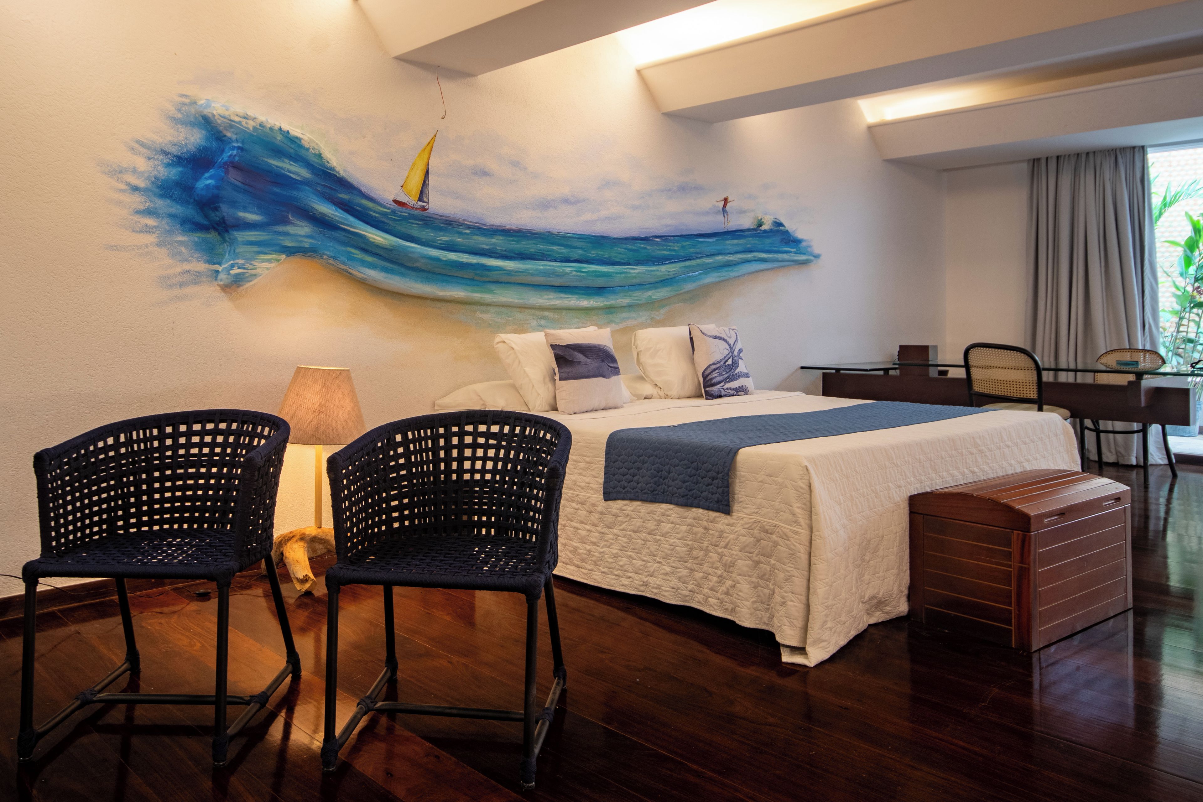 A bedroom with a bed, two chairs, a desk, and a painting of a wave.
