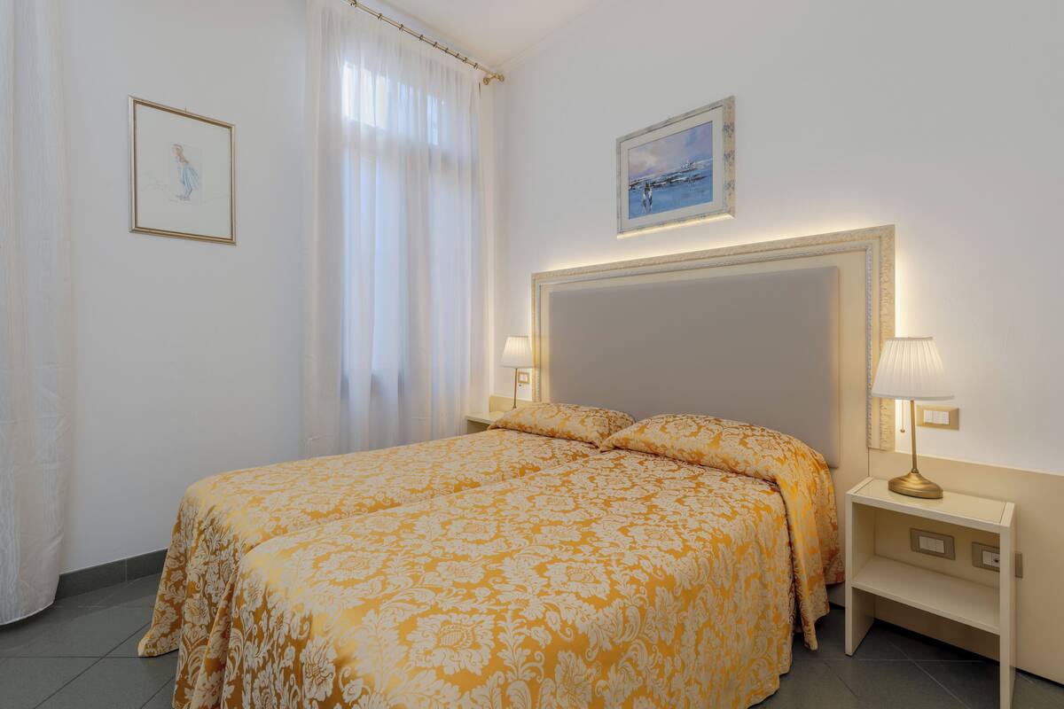 Hotellihuone, jossa on suuri sänky, kaksi yöpöytää, yölamppu, ikkuna verhoilla ja kaksi kehystettyä kuvaa seinällä.