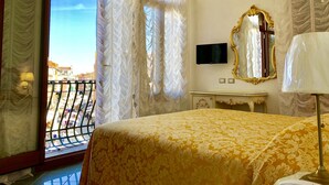 Ein Hotelzimmer mit Bett, Fernseher, Schminktisch und Balkon mit Blick auf Gebäude.