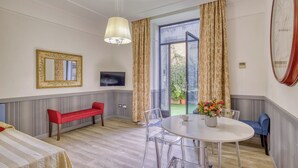 A room with a bed, a red bench, a round table with chairs, a TV, and a large window with curtains. - Lemon Garden (Rome)