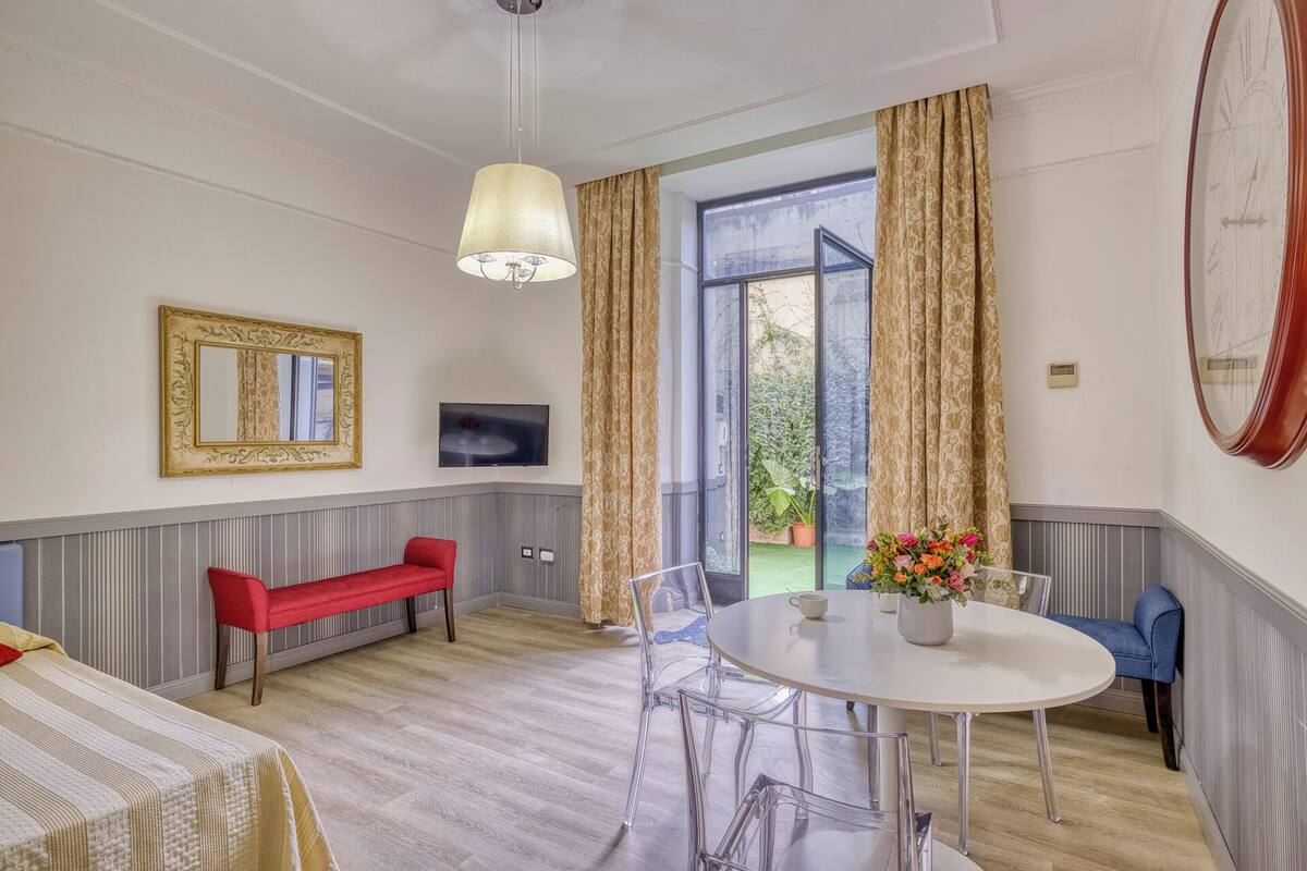 Una habitación con una cama, un banco rojo, una mesa redonda con sillas, un televisor y un ventanal con cortinas.