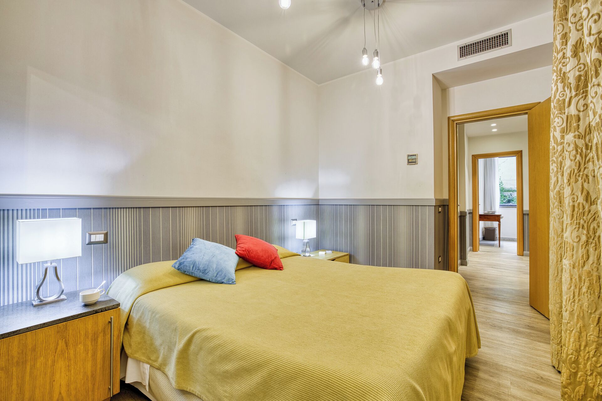 Een hotelkamer met een bed, nachtkastjes, een lamp en uitzicht op de hal.