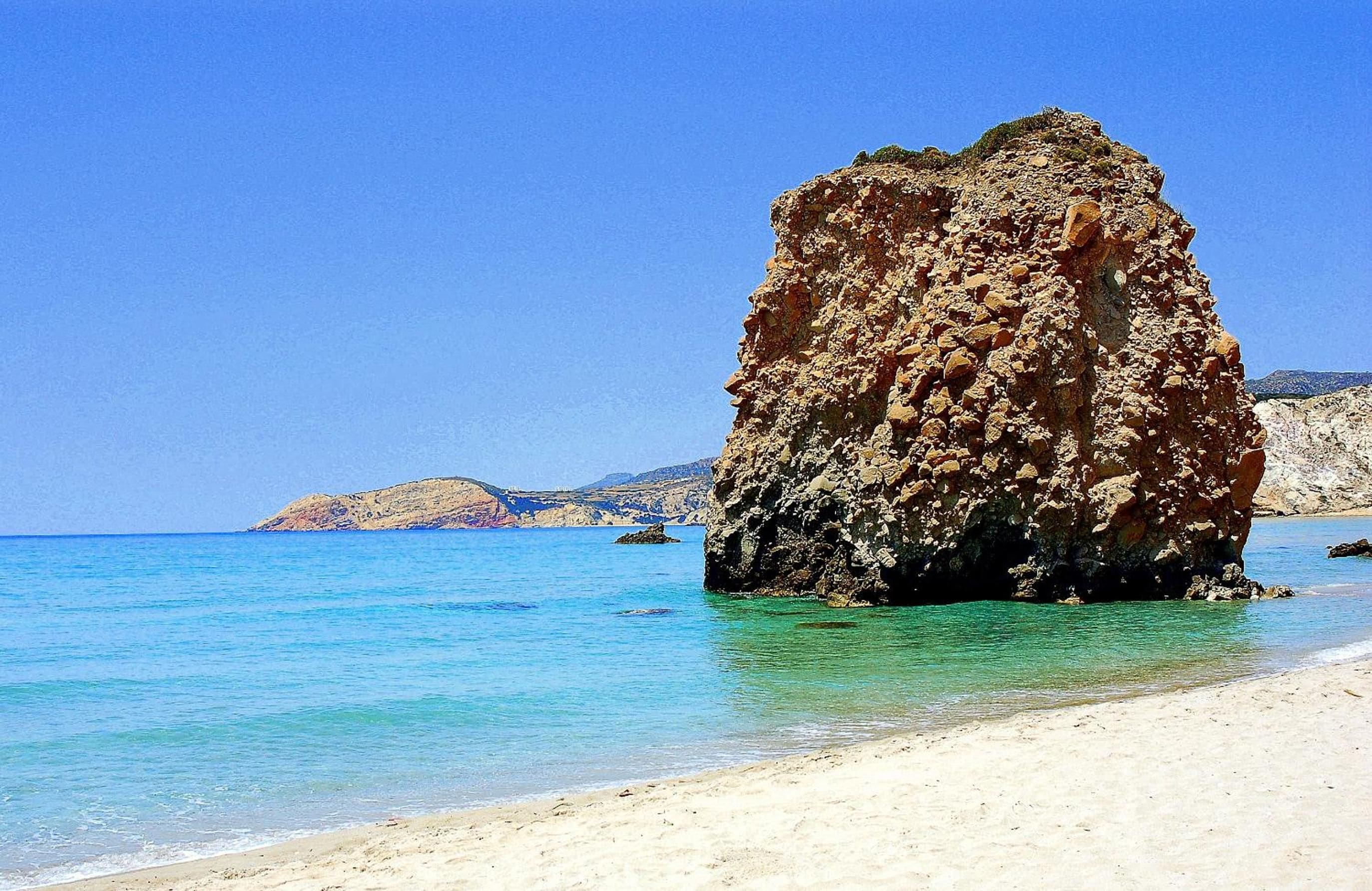 A clear blue sea with a rocky outcrop in the foreground and a sandy beach.