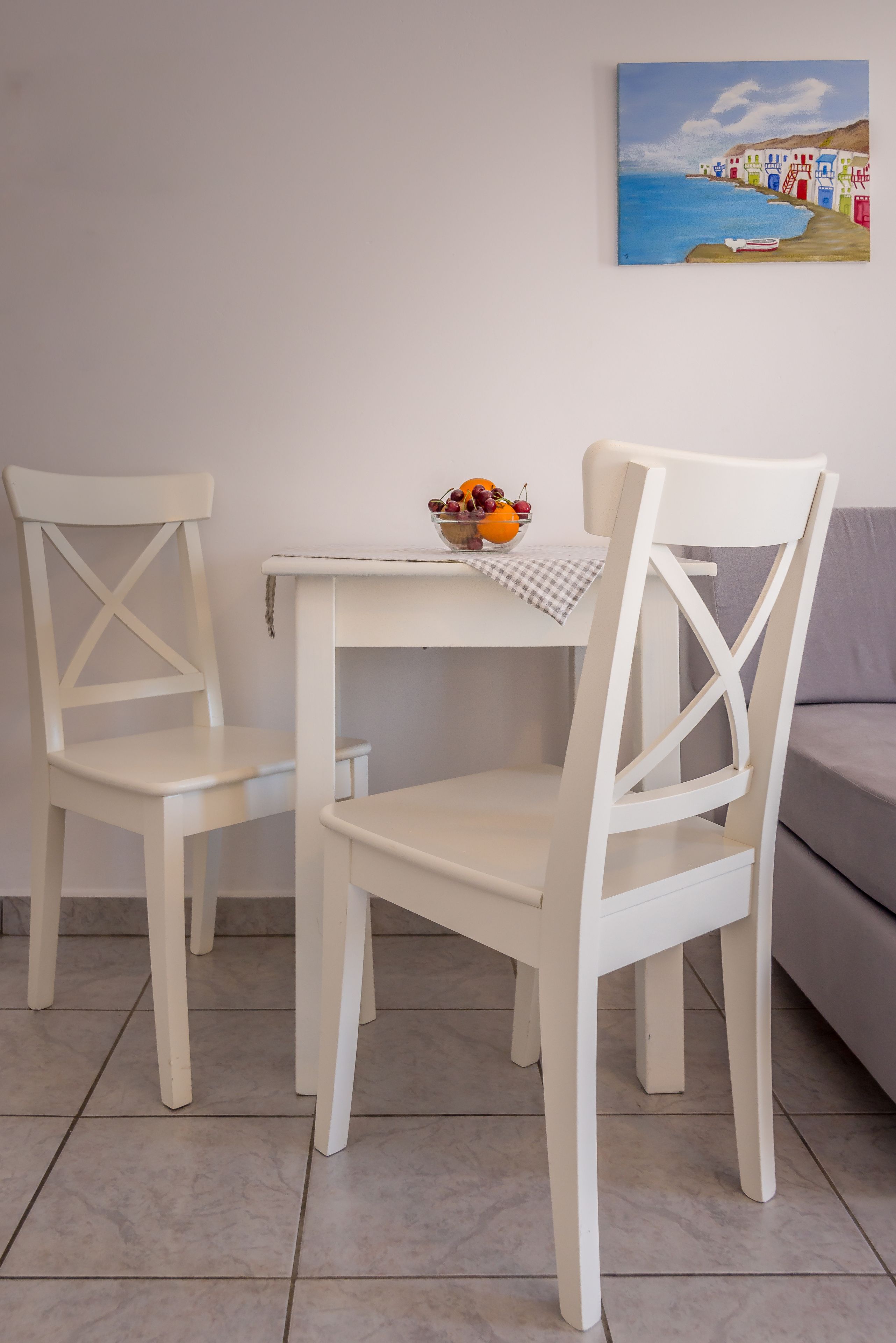 Una mesa de comedor blanca con dos sillas blancas, un cuenco de fruta y un cuadro de una escena costera en la pared.