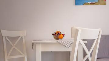 Una mesa de comedor blanca con dos sillas blancas, un cuenco de fruta y un cuadro de una escena costera en la pared.