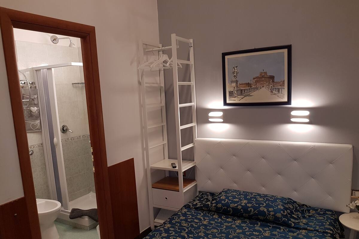 A bedroom with a bed, a white headboard, a shelf unit, a framed picture, and a shower area.