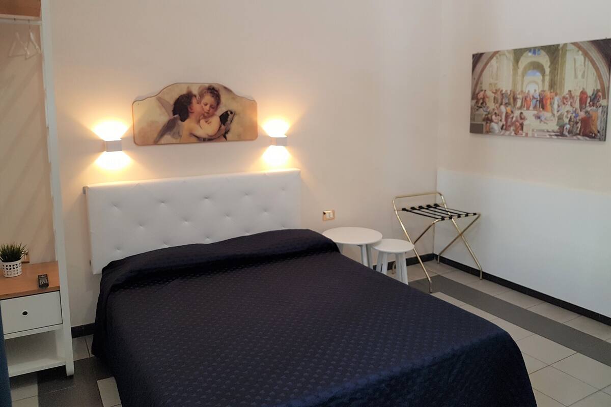A hotel room with a large bed, a small bedside table, a wall-mounted lamp, a painting, and a drying rack.