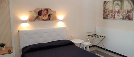 A hotel room with a large bed, a small bedside table, a wall-mounted lamp, a painting, and a drying rack.
