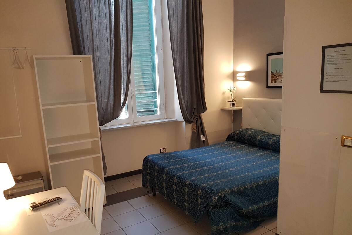 A bedroom with a bed, a desk, a chair, a window with curtains, a bedside lamp, and a framed certificate on the wall.