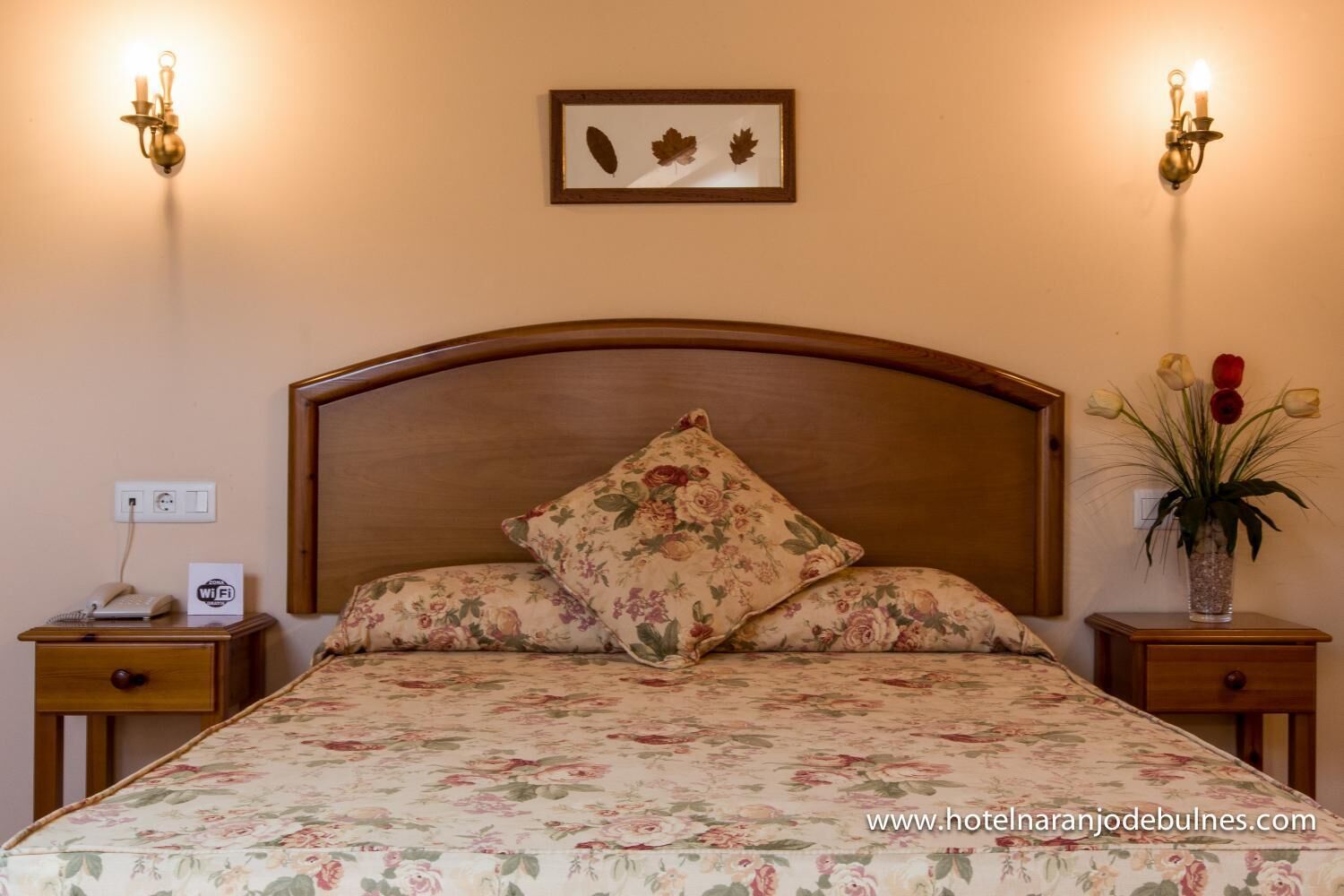 Una cama bien tendida con ropa de cama floral, un cabecero de madera y un cuadro enmarcado encima. Hay dos mesitas de noche con lámparas y un jarrón de flores en una de ellas.