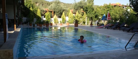 A swimming pool with a child in the water, surrounded by potted plants and lounge chairs.