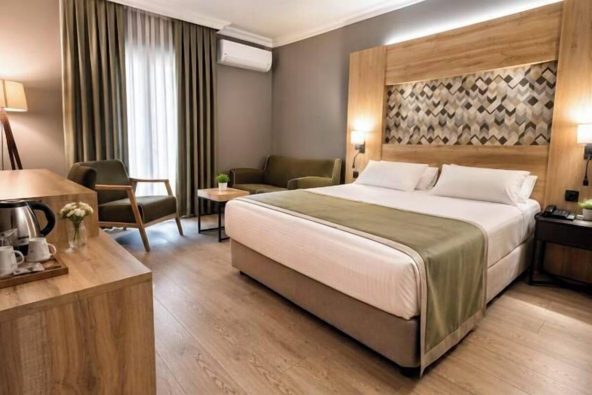 A hotel room with a bed, a desk with a coffee maker, a chair, a small table with a plant, and a wall with a geometric pattern.
