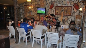 Un grupo de personas cenando en un restaurante con paredes de piedra, sillas de plástico blancas y un televisor montado en la pared.