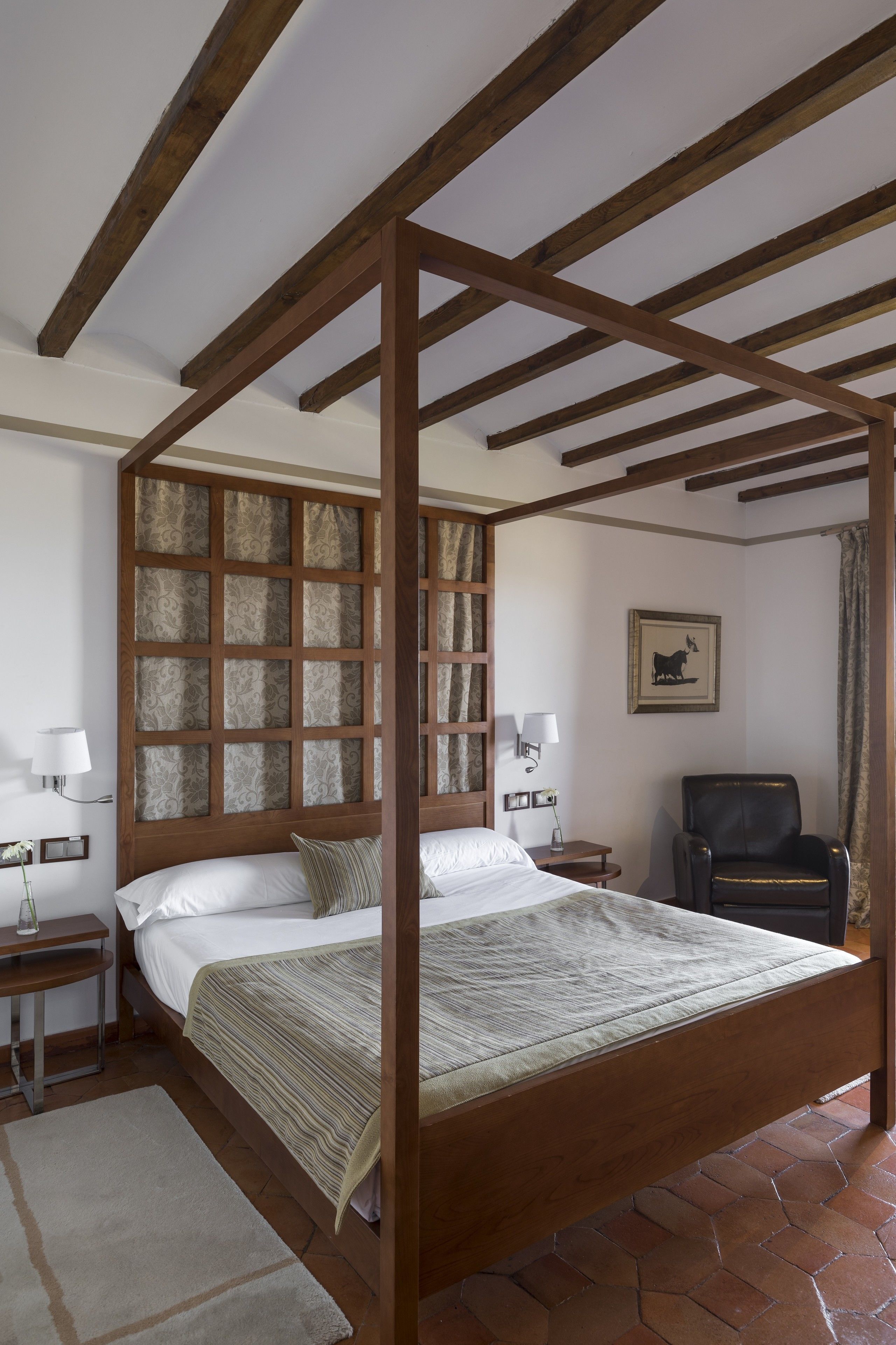 Una cama con dosel de madera, una mesita de noche con lámpara, una silla y una pared con un cuadro enmarcado.