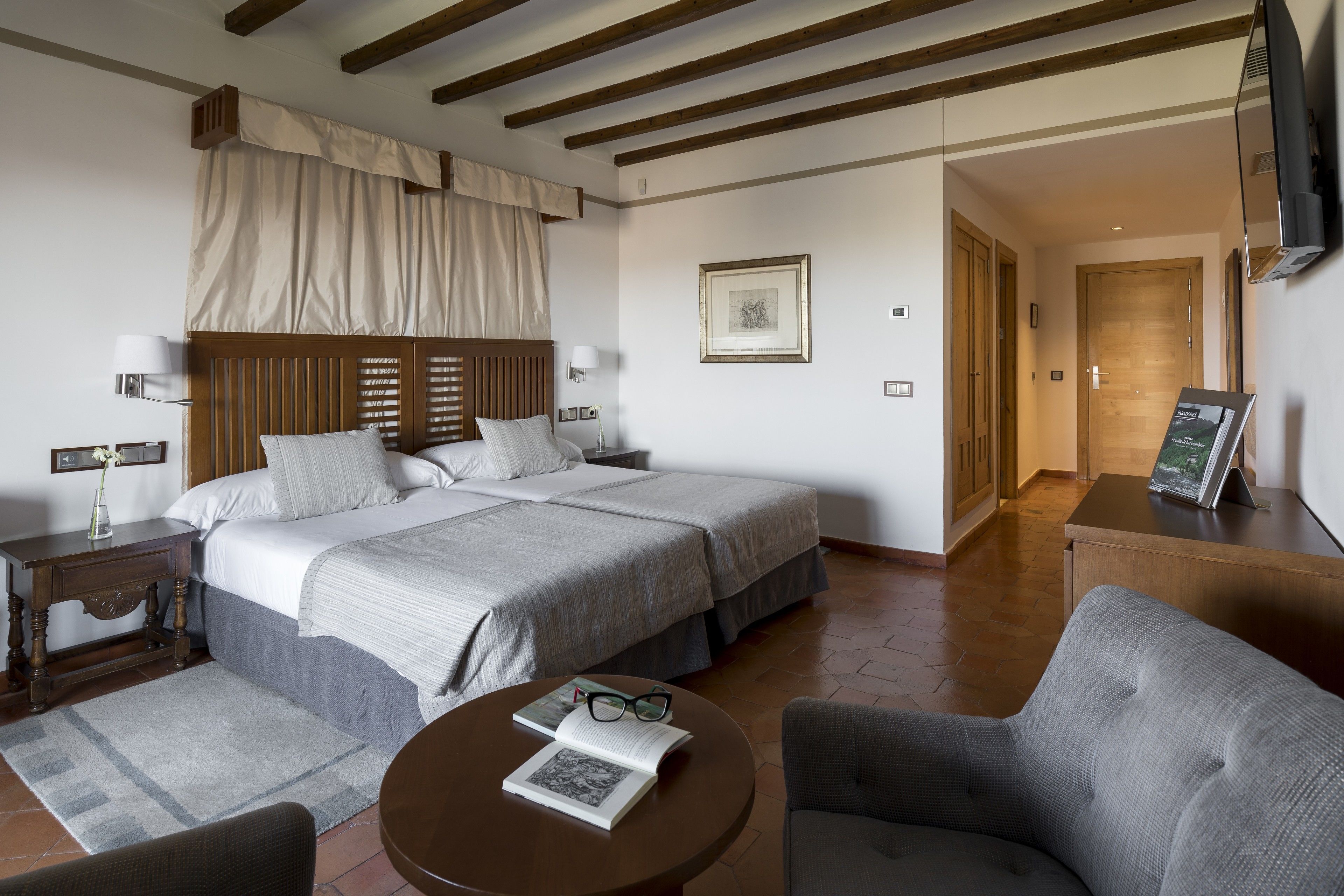 Una camera d'albergo con un letto grande, un comodino con una lampada, un tavolino rotondo con un libro e dei bicchieri, un divano e una TV montata a parete.