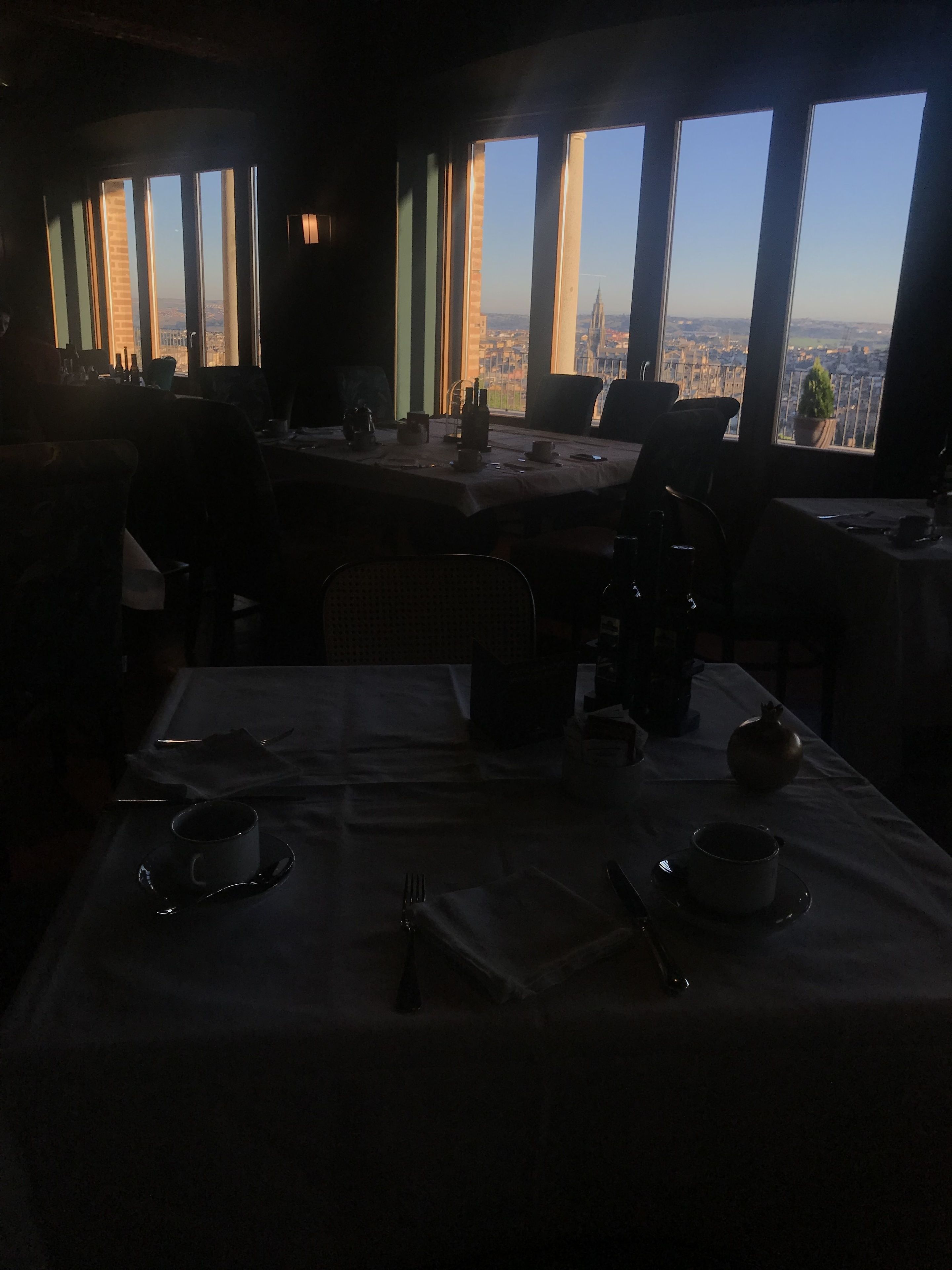Un comedor con poca luz y una mesa dispuesta para dos, con vistas a un paisaje urbano a través de amplias ventanas.