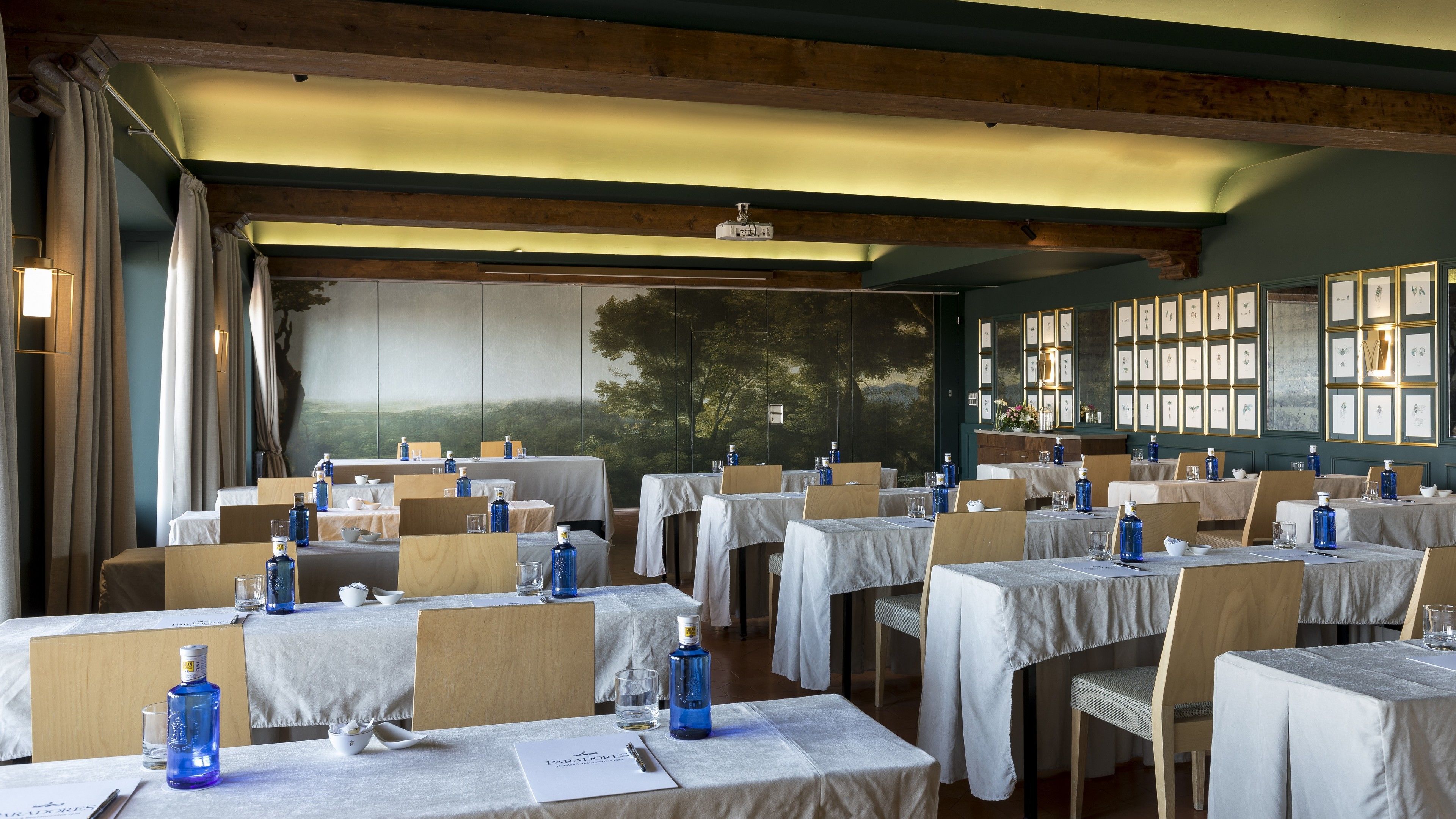 A restaurant with multiple tables set for dining, featuring a mural of trees and a framed picture collection on the wall.