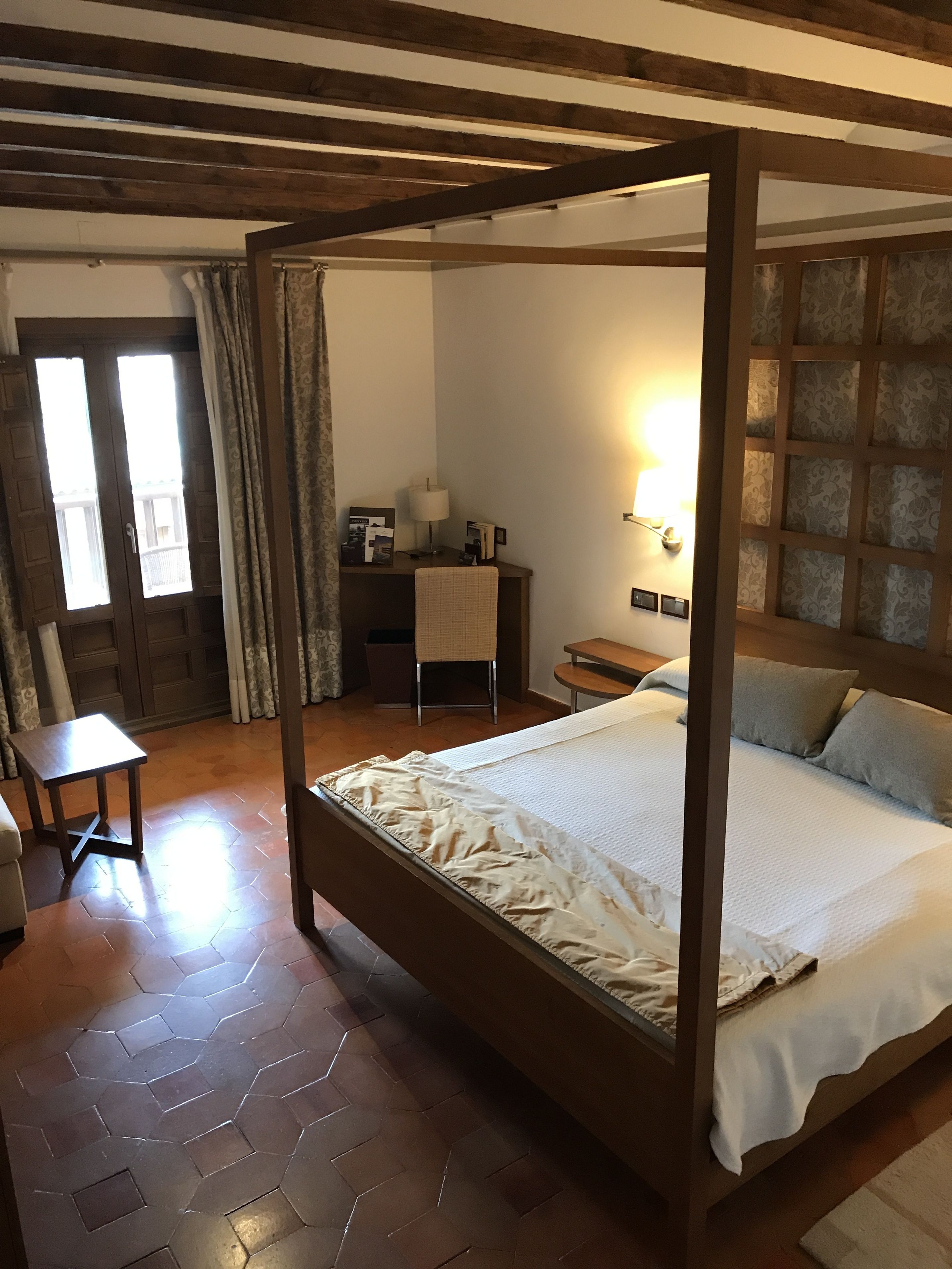 A four-poster bed with a white bedspread and two pillows, a wooden desk with a lamp, a chair, and a wall-mounted light fixture.