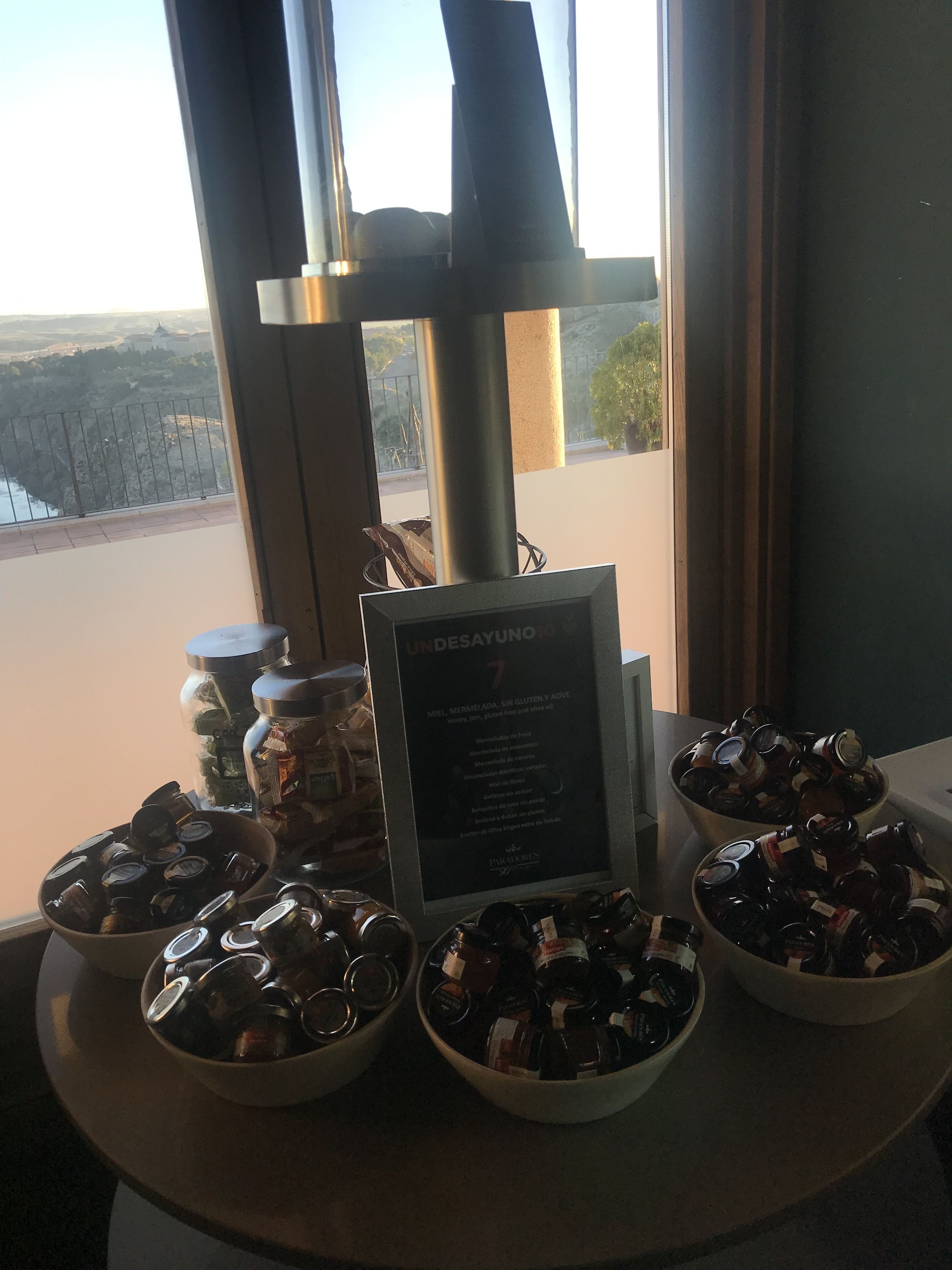 Ett bord med burkar fulla av honung och en menytavla med texten ”UNDESAYUNOS 7”, med utsikt över ett bergigt landskap genom stora fönster.