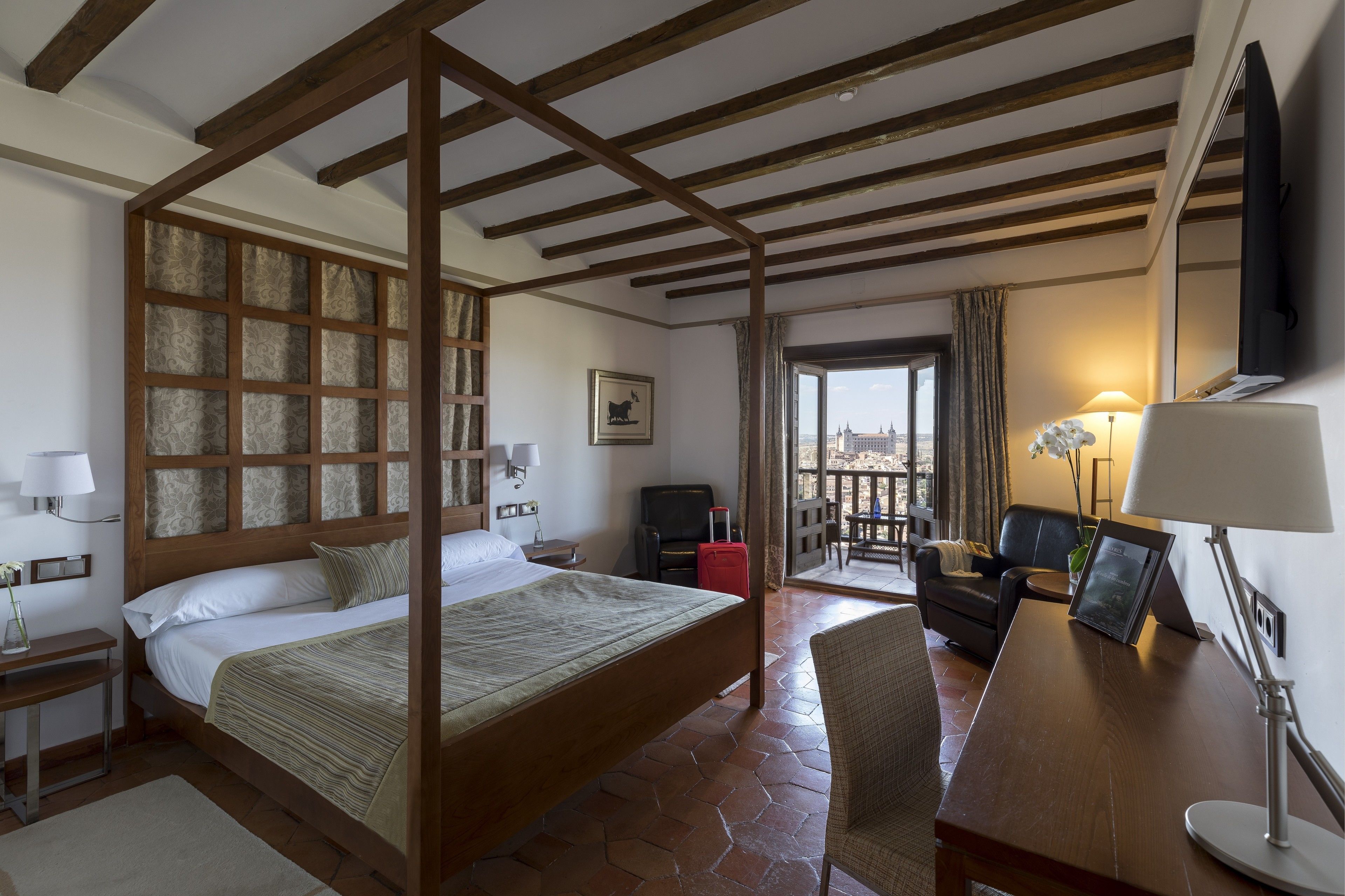 Een hotelkamer met een houten hemelbed, een bureau met lamp, een stoel, een televisie en uitzicht op de stad door een raam.