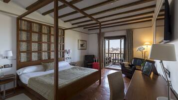 Een hotelkamer met een houten hemelbed, een bureau met lamp, een stoel, een televisie en uitzicht op de stad door een raam.