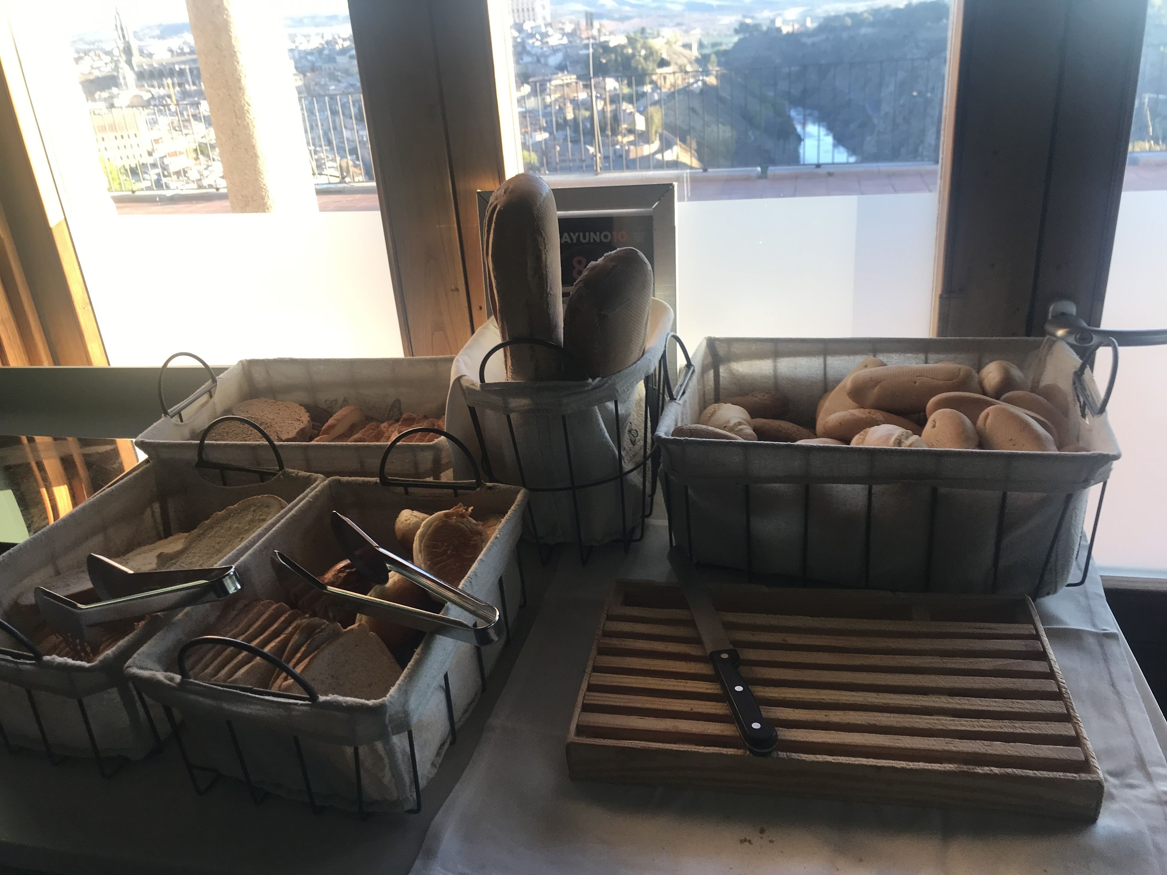 A breakfast buffet with various bread and pastries in wire baskets, a knife on a cutting board, and a view of a cityscape through large windows.