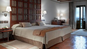 Habitación de hotel con una cama grande, cabecera de madera, dos mesitas de noche con lámparas, un escritorio con una silla y vistas a través de un ventanal.