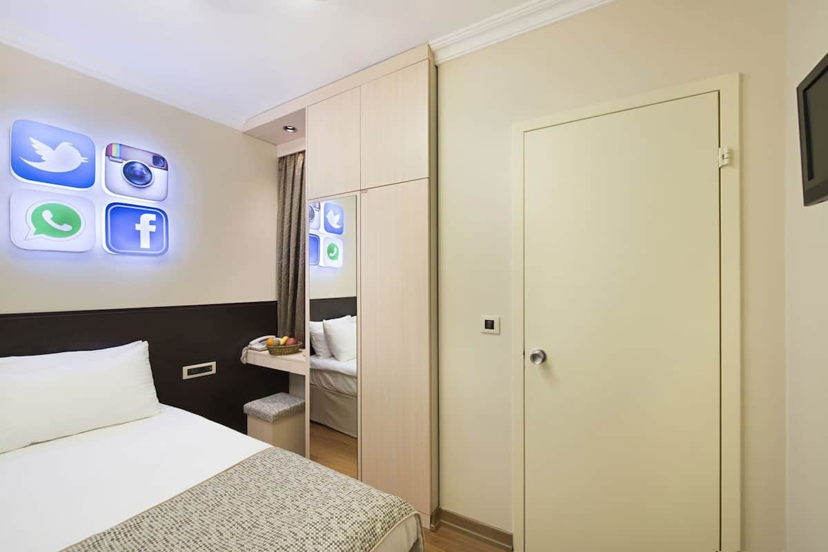 Habitación de hotel con cama, armario, un pequeño escritorio con una cesta de fruta e iconos de redes sociales iluminados en la pared.