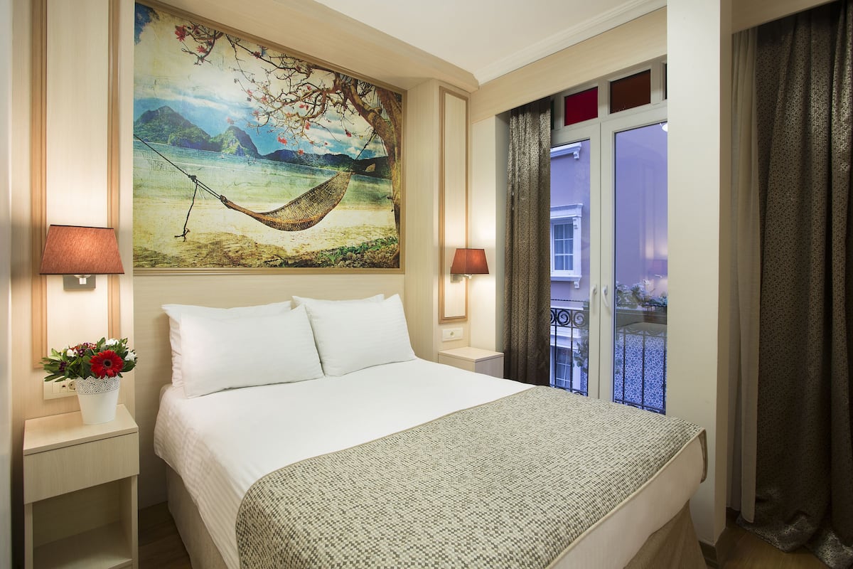 Una habitación de hotel con una cama grande, dos mesitas de noche, una mesita de noche con un jarrón de flores, un cuadro de una hamaca y vistas al exterior a través de una puerta corredera.