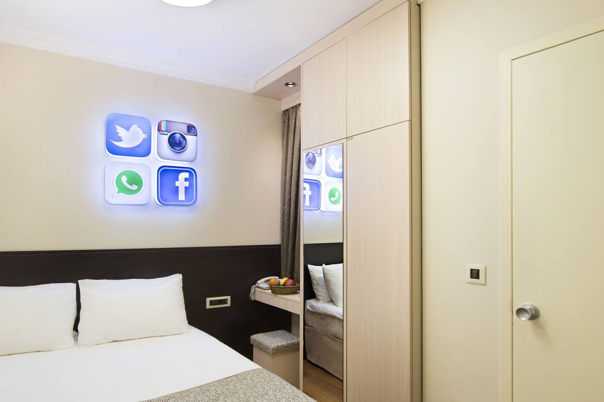 Habitación de hotel con cama, mesita de noche, armario y una pared con iconos de redes sociales iluminados.