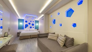 A modern hotel room with a bed, a sofa, a desk, and a wall with geometric light fixtures.