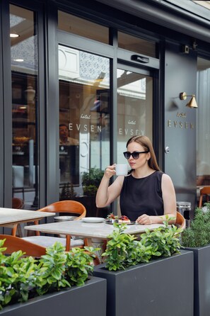 A person seated at an outdoor cafe with a table and chairs, holding a cup. The cafe has a modern design with glass doors and windows. - Hotel Evsen - Adults Only (Istanbul)