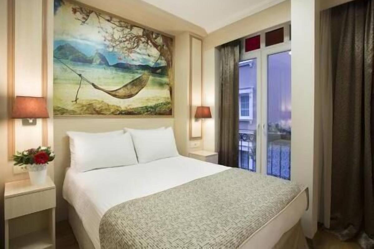 A hotel room with a bed, two bedside lamps, a painting, a window with curtains, and a balcony view.
