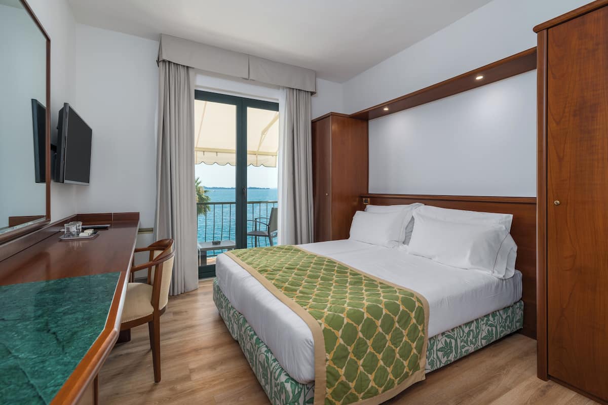 A hotel room with a bed, a desk with a chair, a television, and a balcony with a view of the sea.
