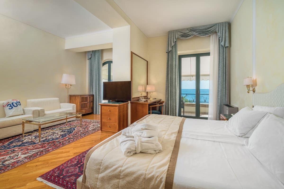 A hotel room with a bed, a sofa, a desk, a television, and a view of the sea.