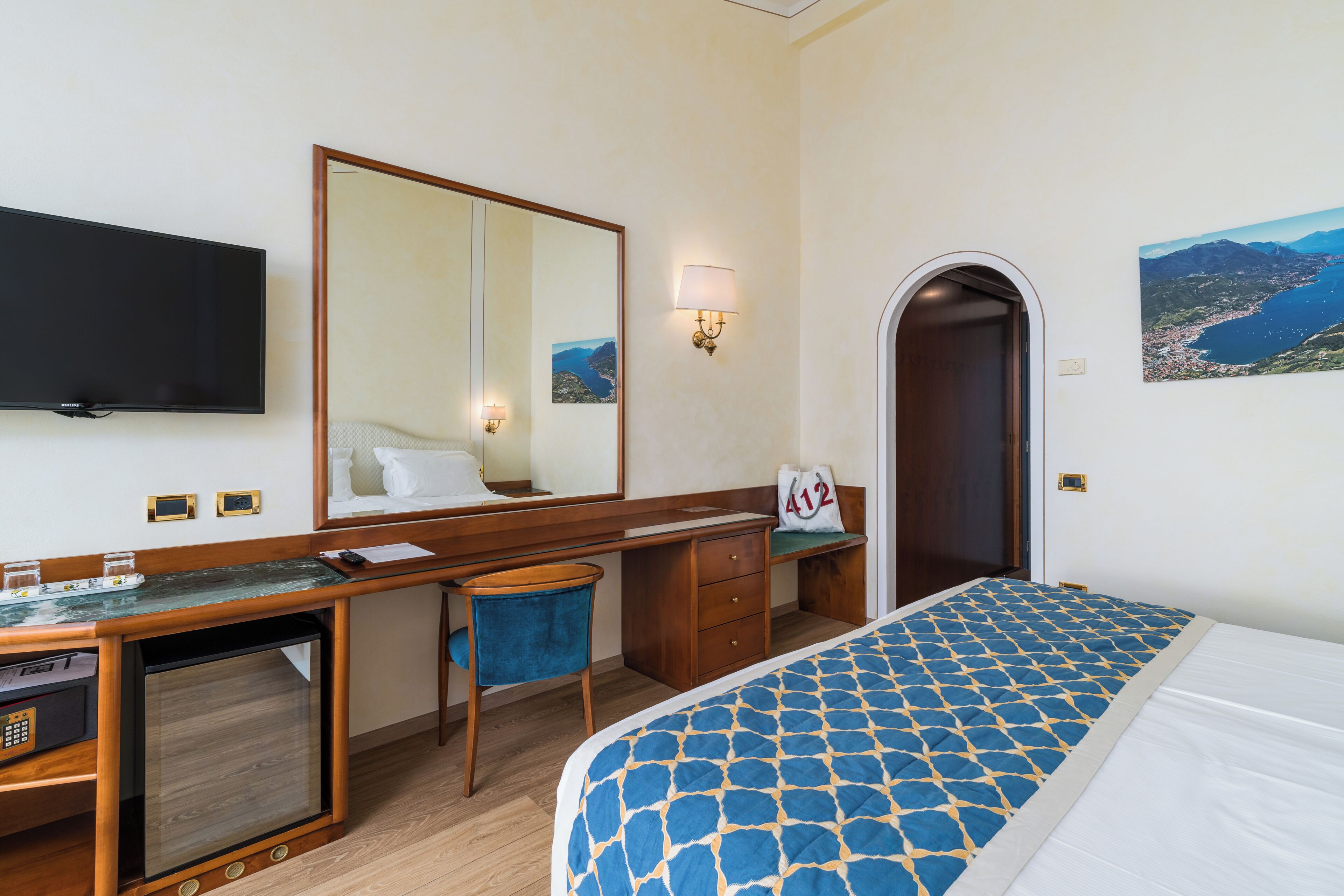 Quarto de hotel com cama, escrivaninha, cadeira, televisão e porta que dá para outro cômodo.