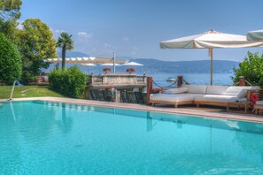 A swimming pool with lounge chairs and umbrellas, a table with flowers, and a view of the sea and mountains. - Villa Capri (Gardone Riviera)