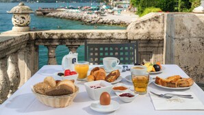 A breakfast table with croissants, bread, fruit, and beverages, overlooking a coastal area with a tower and boats. - Villa Capri (Gardone Riviera)