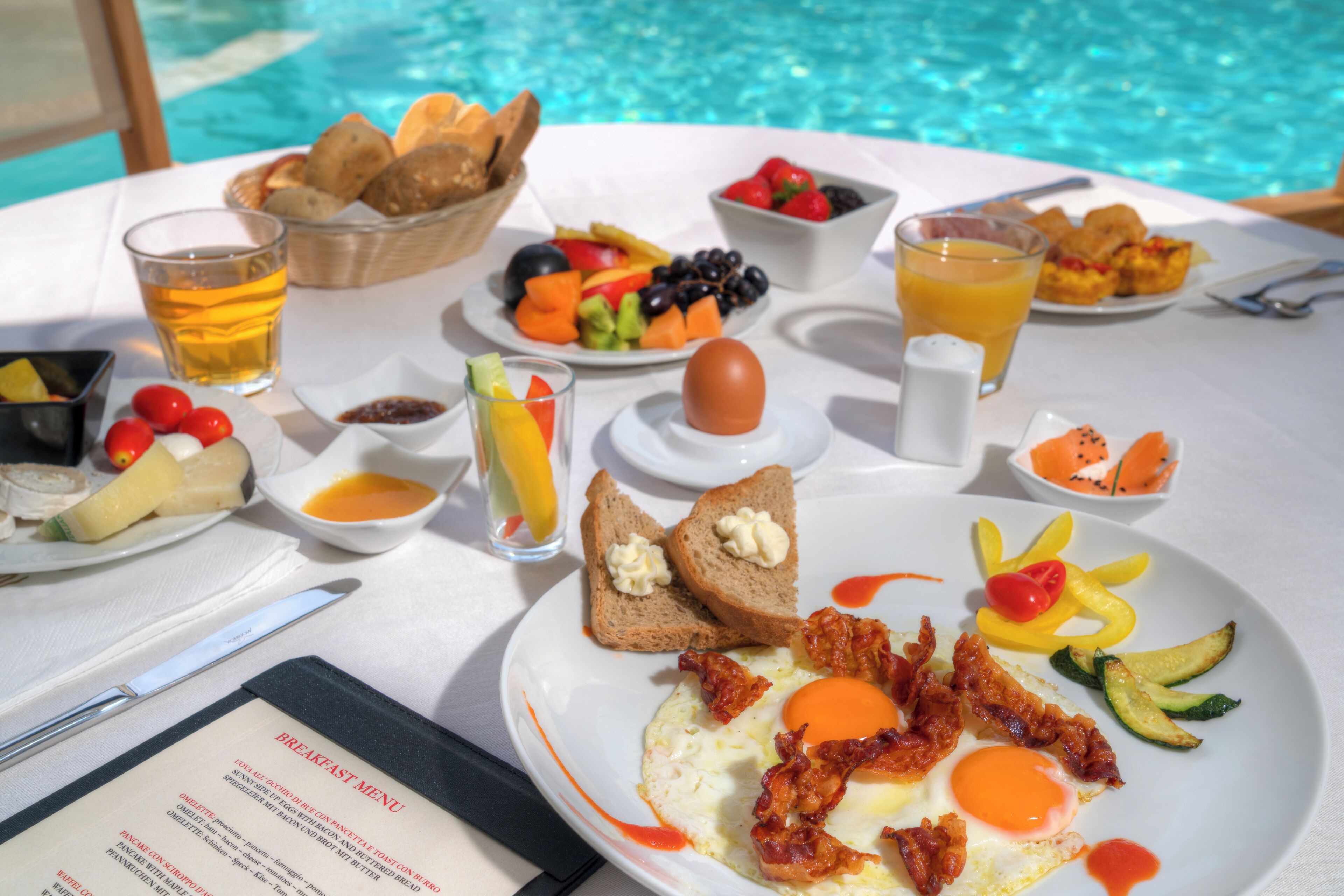A breakfast buffet with a variety of fruits, vegetables, eggs, and toast by a poolside table.