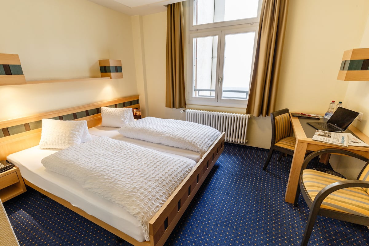 Ein Hotelzimmer mit Doppelbett, Schreibtisch und Stuhl, Heizkörper und einem Fenster mit Vorhängen.