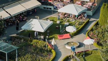 An outdoor dining area with tables and umbrellas, surrounded by a garden with hedges and flowers.